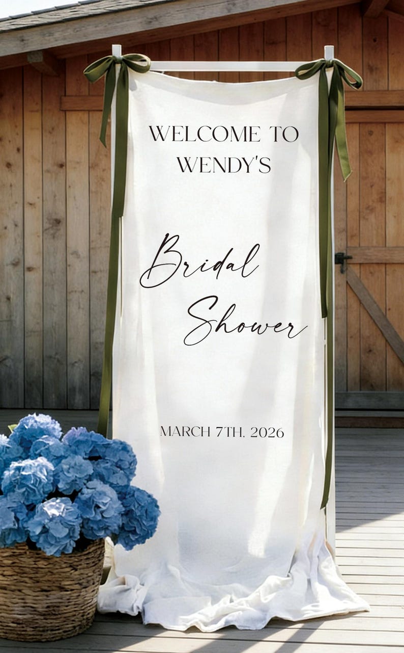 May include: A white fabric banner with the text "WELCOME TO WENDY'S Bridal Shower" and the date "MARCH 7TH, 2026". The banner is adorned with green ribbons and a basket of blue flowers.