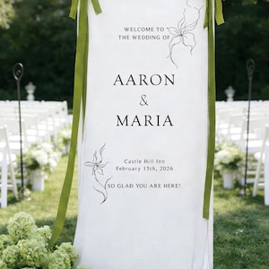 May include: A white wedding sign with green ribbons and floral accents. The sign reads "Welcome to the Wedding of Aaron & Maria" with the date and location. The sign is set up outdoors with white chairs in the background.