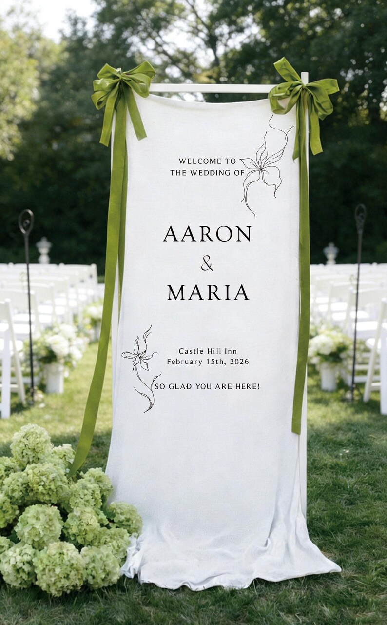 May include: A white wedding sign with green ribbons and floral accents. The sign reads "Welcome to the Wedding of Aaron & Maria" with the date and location. The sign is set up outdoors with white chairs in the background.