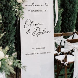 May include: A white wedding welcome sign with black script that reads "welcome to the wedding of Olivia & Dylan, July 15th, 2025, We are so glad you are here!" The sign is decorated with white flowers and greenery.