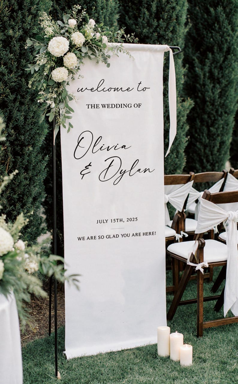 May include: A white wedding welcome sign with black script that reads "welcome to the wedding of Olivia & Dylan, July 15th, 2025, We are so glad you are here!" The sign is decorated with white flowers and greenery.