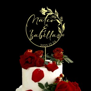 K&ouml;nnte beinhalten: Goldener Hochzeitstortenaufsatz mit den Namen Mateo & Isabella und dem Datum 2025.12.24. Der Aufsatz befindet sich auf einer wei&szlig;en Torte, die mit roten Rosenbl&uuml;ten verziert ist.
