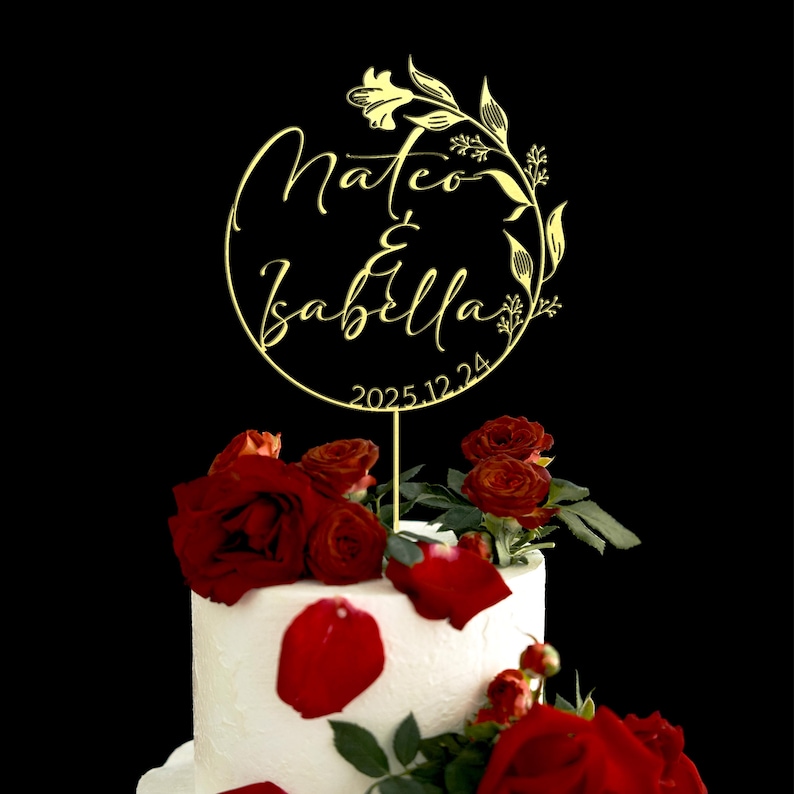 K&ouml;nnte beinhalten: Goldener Hochzeitstortenaufsatz mit den Namen Mateo & Isabella und dem Datum 2025.12.24. Der Aufsatz befindet sich auf einer wei&szlig;en Torte, die mit roten Rosenbl&uuml;ten verziert ist.
