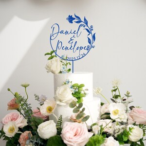 K&ouml;nnte beinhalten: Ein blauer Tortendekor mit floralem Design und dem Text "Daniel & Penelope 2025.12.24" auf einem wei&szlig;en Kuchen.