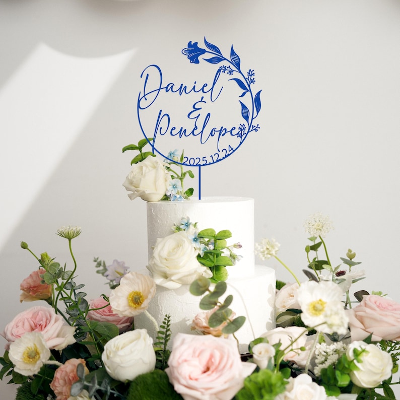 K&ouml;nnte beinhalten: Ein blauer Tortendekor mit floralem Design und dem Text "Daniel & Penelope 2025.12.24" auf einem wei&szlig;en Kuchen.
