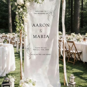 May include: A white wedding welcome sign with the names "Aaron & Maria" and the date February 15th, 2026. The sign is decorated with floral arrangements and set in a wooded area. The sign reads "Welcome to the wedding of... So glad you are here!"