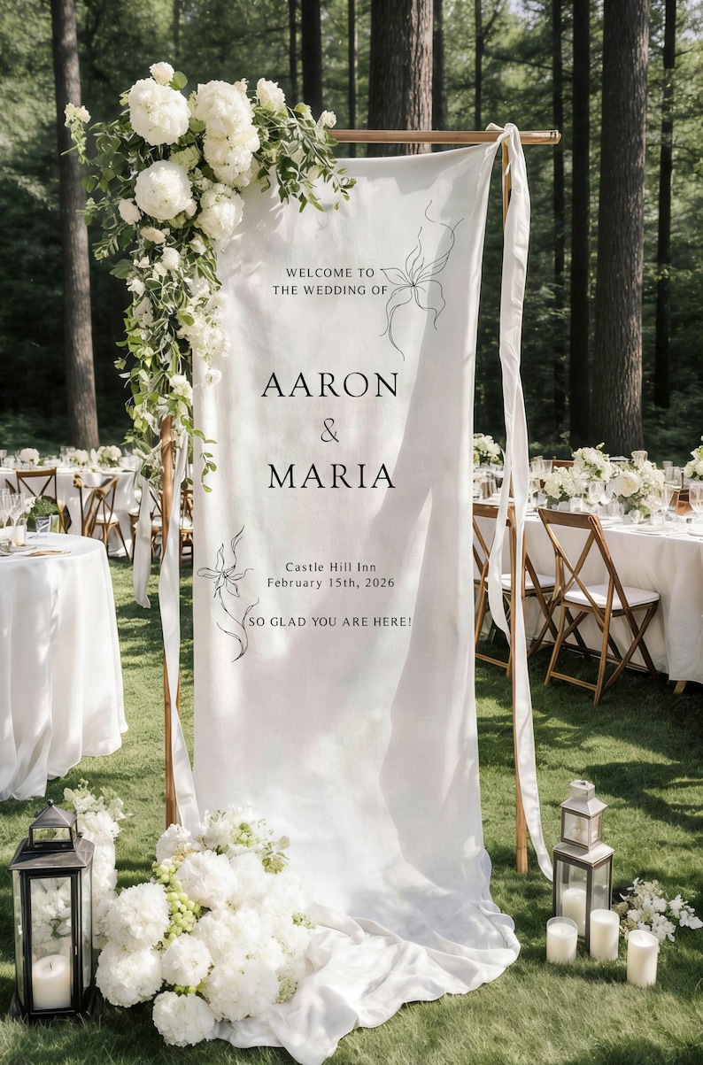 May include: A white wedding welcome sign with the names "Aaron & Maria" and the date February 15th, 2026. The sign is decorated with floral arrangements and set in a wooded area. The sign reads "Welcome to the wedding of... So glad you are here!"