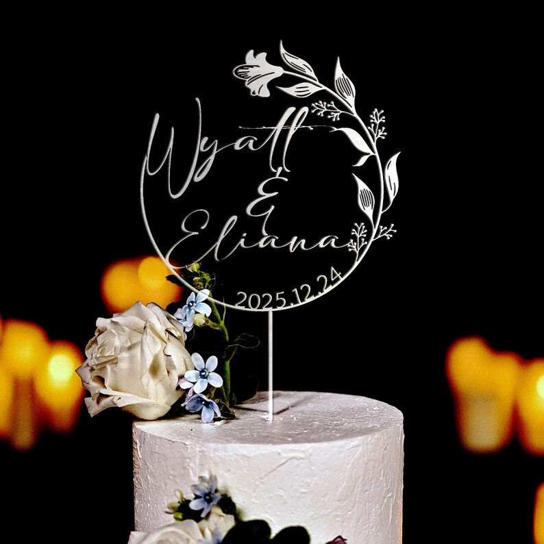 K&ouml;nnte beinhalten: Wei&szlig;er Acryl-Kuchentopper mit einem runden Design mit den Namen Wyatt & Eliana und dem Datum 2025.12.24. Der Topper ist mit floralen Designs verziert.