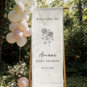 May include: A baby shower sign with the text "Welcome to Ariane Baby Shower 08.02.2026" and a floral illustration. The sign is on a wooden frame, with white and pink balloons and floral arrangements. The background is a natural outdoor setting.