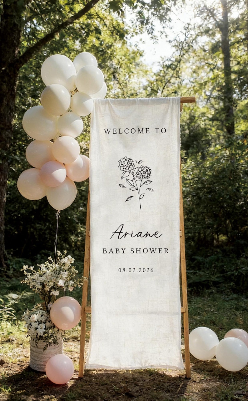 May include: A baby shower sign with the text "Welcome to Ariane Baby Shower 08.02.2026" and a floral illustration. The sign is on a wooden frame, with white and pink balloons and floral arrangements. The background is a natural outdoor setting.