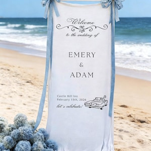 May include: A white wedding sign on a beach with the text "Welcome to the wedding of Emery & Adam". The sign includes the date, location, and a car illustration. Light blue bows and ribbons adorn the top.