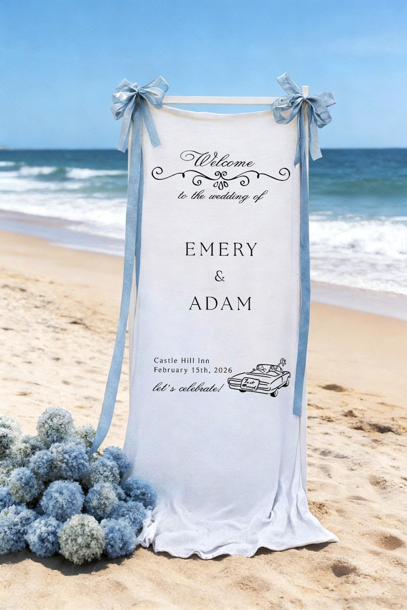 May include: A white wedding sign on a beach with the text "Welcome to the wedding of Emery & Adam". The sign includes the date, location, and a car illustration. Light blue bows and ribbons adorn the top.