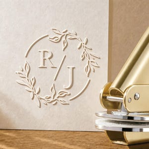 以下が含まれることがあります： 金色の金属製エンボス加工ツールと、クリーム色の紙。紙には、繊細で様式化された葉の枝と茎で構成された円形のフレームの中に、文字「R」と「J」が中央に配置された浮き彫りのエンボスデザインが施されています。