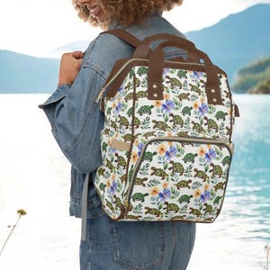 May include: A white backpack with a pattern of green turtles, yellow and purple flowers, and green leaves. The backpack has brown trim, straps, and handles. It has a front pocket and is being worn outdoors.