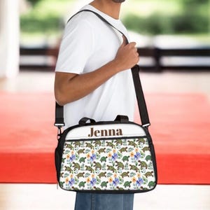 May include: A black and white shoulder bag with a turtle and floral pattern. The bag has a black strap and the name "Jenna" printed on the top. The bag is being carried by a person wearing a white t-shirt.