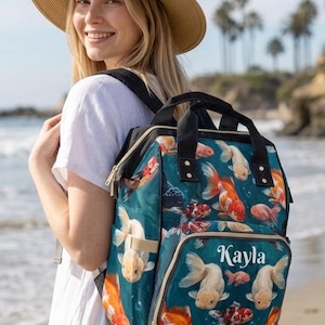 May include: A teal backpack featuring a repeating pattern of orange, white, and red goldfish. The bag has black straps, gold-tone zippers, and a front pocket with the name Kayla printed in white script lettering across the center of the pocket.