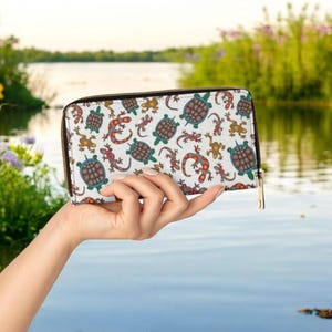 May include: A white wallet with a colorful pattern of turtles and lizards. The wallet has a black zipper and a gold-colored zipper pull. The wallet is held in front of a blurred background of water and greenery.