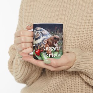 Pode incluir: Caneca de cerâmica branca com uma imagem colorida de uma tartaruga leopardo com uma flecha atravessando um coração. O texto "Eu amo tartarugas leopardo" está impresso na caneca.