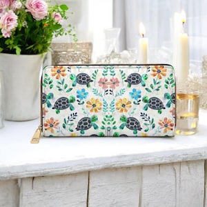 May include: A white wallet with a colorful pattern of turtles, flowers, and leaves. The wallet has a gold zipper and a rectangular shape. The design includes orange, blue, and green elements.