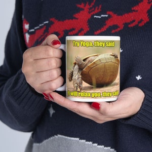 Pode incluir: Caneca de cerâmica branca com uma tartaruga em pose de ioga. A caneca tem o texto "Try Yoga, they said" e "it will relax you, they said" em amarelo. A caneca é segurada por uma pessoa que usa um suéter azul marinho.