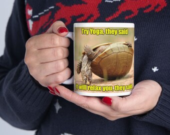 Może przedstawiać: Biały ceramiczny kubek z żółwiem w pozycji jogi. Kubek ma tekst "Try Yoga, they said" i "it will relax you, they said" w żółtym kolorze. Kubek jest trzymany przez osobę w granatowym swetrze.