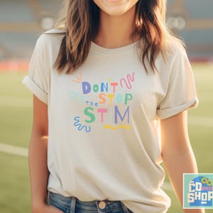 Pu&ograve; includere: Una maglietta bianca con il testo "Don't Stop the Stim" in lettere e vortici colorati. La maglietta &egrave; indossata da una persona con i capelli lunghi e castani.