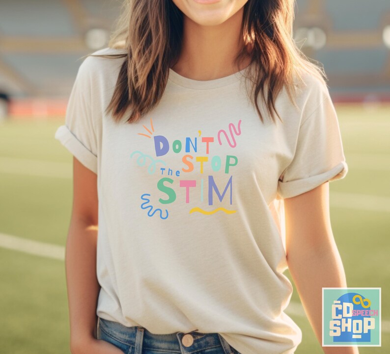 Pu&ograve; includere: Una maglietta bianca con il testo "Don't Stop the Stim" in lettere e vortici colorati. La maglietta &egrave; indossata da una persona con i capelli lunghi e castani.