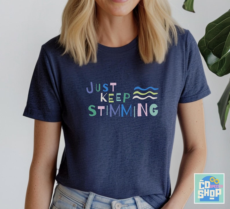 May include: A navy blue t-shirt with the text 'JUST KEEP STIMMING' in colorful letters. The text is in a playful font and includes a wavy line design.