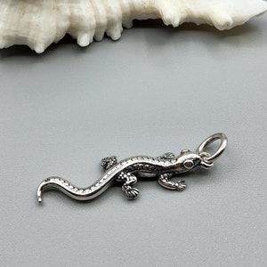 May include: A silver lizard charm with a textured surface. The charm has a loop at the top for attaching to a necklace or bracelet.