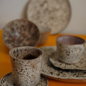 May include: A set of ceramic tableware featuring two cylindrical cups and two matching saucers. The pieces have a speckled, off-white and brown textured finish. In the background, an additional bowl and a large plate are visible.
