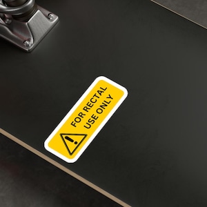 Puede incluir: Una tabla de skate negra con una pegatina amarilla de advertencia que dice "FOR RECTAL USE ONLY". La pegatina tiene un signo de exclamación negro. La tabla de skate tiene ruedas rojas y azules.