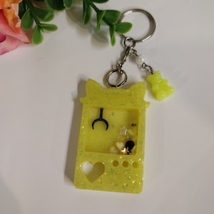 May include: A yellow keychain shaped like a handheld video game with glitter embedded in the resin. The keychain has a black crescent moon, stars, and hearts inside the game screen. A small yellow gummy bear charm hangs from the keychain.