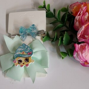 May include: Two hair clips with bows. One is a light blue bow with a cupcake character, and the other is a small, glittery blue bow. Both are attached to silver metal clips. The bows are on white cardstock.