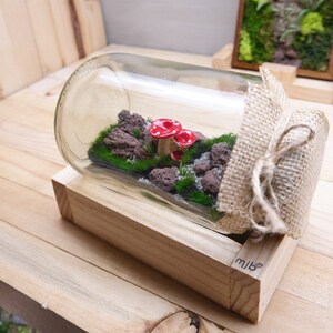 May include: A glass bottle terrarium with a wooden base. The bottle contains red mushrooms, green moss, and small rocks. The bottle is wrapped in burlap and tied with twine. The base is labeled "mibe".