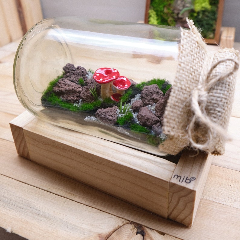May include: A glass jar terrarium with a miniature forest scene. The jar is filled with moss, small rocks, and two red mushroom figurines. The jar is wrapped in burlap and tied with twine. The jar is sitting on a wooden base with the word "mibe" engraved on it.