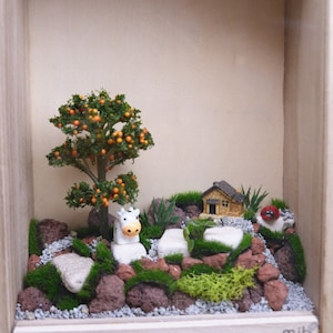May include: A miniature scene in a wooden frame with a small white cow figurine, a miniature house, and a small orange tree. The scene is decorated with moss, rocks, and gravel.