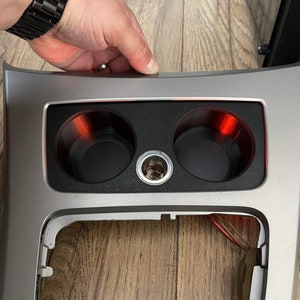 Può includere: Un rivestimento interno in argento per auto con due portabevande e una presa di corrente. I portabevande sono neri e hanno un bagliore rosso intorno.