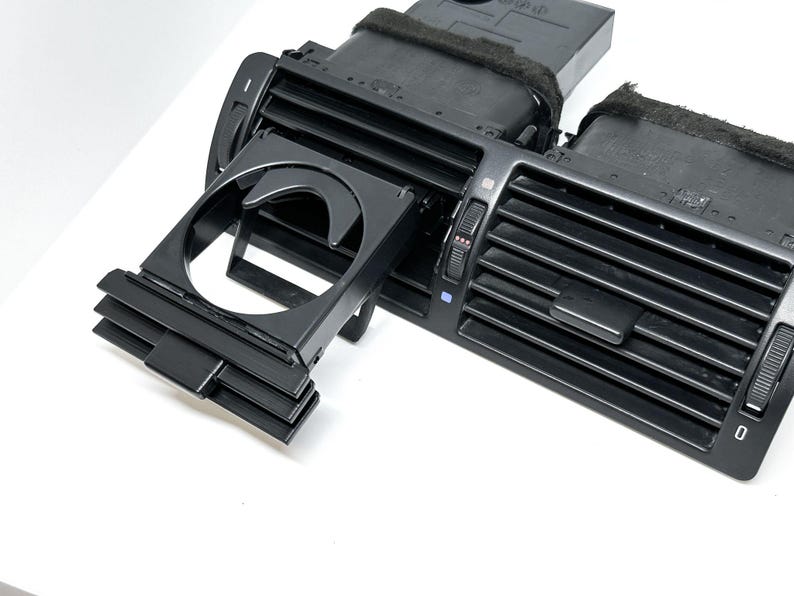 Peut inclure: A&eacute;rateur d'air de voiture en plastique noir avec un porte-gobelet int&eacute;gr&eacute;. L'a&eacute;rateur est dot&eacute; d'une s&eacute;rie de lamelles horizontales et de deux boutons sur le c&ocirc;t&eacute;. Le porte-gobelet est r&eacute;tractable et poss&egrave;de une ouverture circulaire.