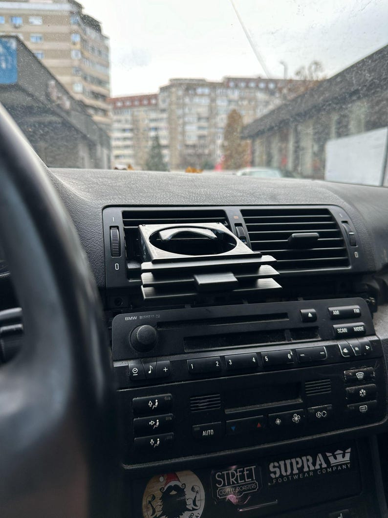 Peut inclure: Un tableau de bord de voiture noir avec un porte-gobelet noir, une radio avec des boutons et un autocollant qui dit "SUPRA FOOTWEAR COMPANY".