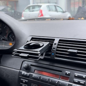 Peut inclure: Porte-gobelet de voiture noir fix&eacute; &agrave; l'a&eacute;rateur de la planche de bord d'une voiture. Le porte-gobelet est en plastique et poss&egrave;de un m&eacute;canisme &agrave; ressort pour maintenir le gobelet en place. La planche de bord de la voiture est noire et poss&egrave;de un affichage num&eacute;rique indiquant la temp&eacute;rature, qui est de 23,0 degr&eacute;s Celsius.