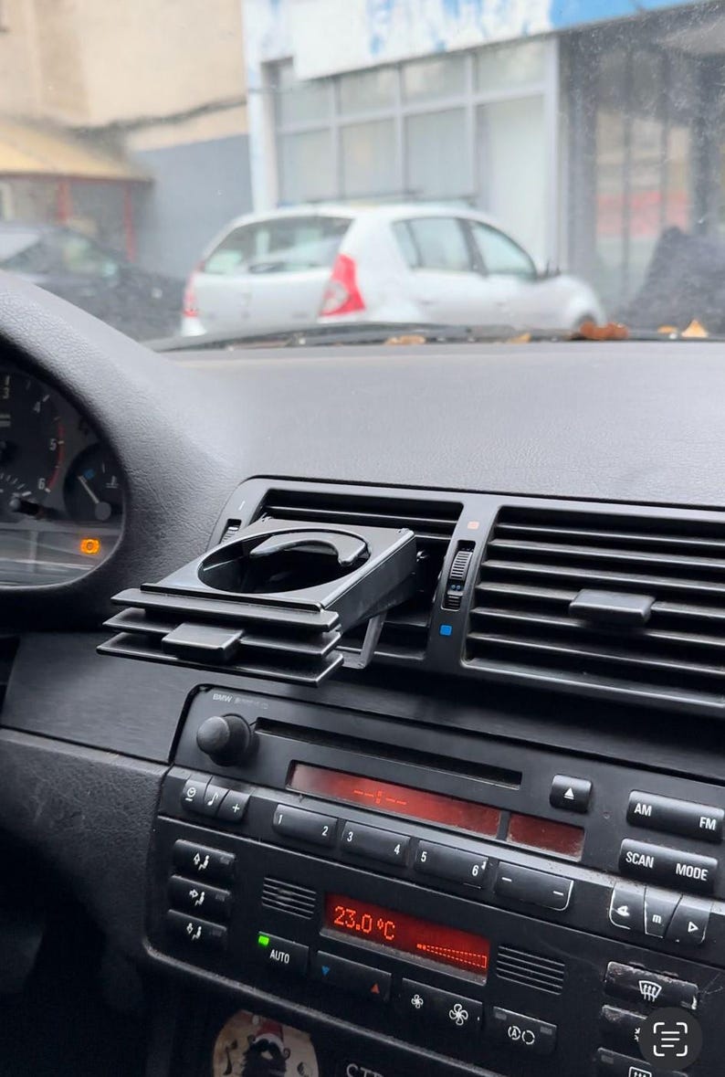 Peut inclure: Porte-gobelet de voiture noir fix&eacute; &agrave; l'a&eacute;rateur de la planche de bord d'une voiture. Le porte-gobelet est en plastique et poss&egrave;de un m&eacute;canisme &agrave; ressort pour maintenir le gobelet en place. La planche de bord de la voiture est noire et poss&egrave;de un affichage num&eacute;rique indiquant la temp&eacute;rature, qui est de 23,0 degr&eacute;s Celsius.