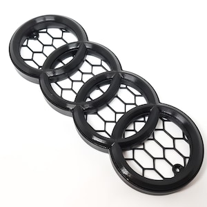 Op de afbeelding: Zwart plastic Audi-embleem met vier in elkaar grijpende ringen. De ringen hebben een honingraatpatroon.