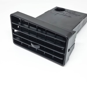 Può includere: Bocchetta di ventilazione per auto in plastica nera con una griglia rettangolare e una piccola manopola rotonda sul lato. La bocchetta è progettata per dirigere il flusso d'aria dal sistema di climatizzazione dell'auto.