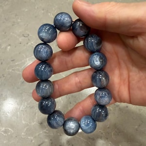 May include: A stretch bracelet composed of fourteen large, round, polished kyanite beads. The beads exhibit a deep blue colour with characteristic fibrous, chatoyant inclusions and subtle greyish-white streaks throughout the stone surfaces.