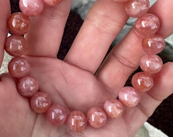 May include: A bracelet made of round, translucent, pink and orange gemstone beads. The beads are strung together, forming a circular shape. The bracelet is held in a hand, showcasing the natural variations in color and texture of the stones.
