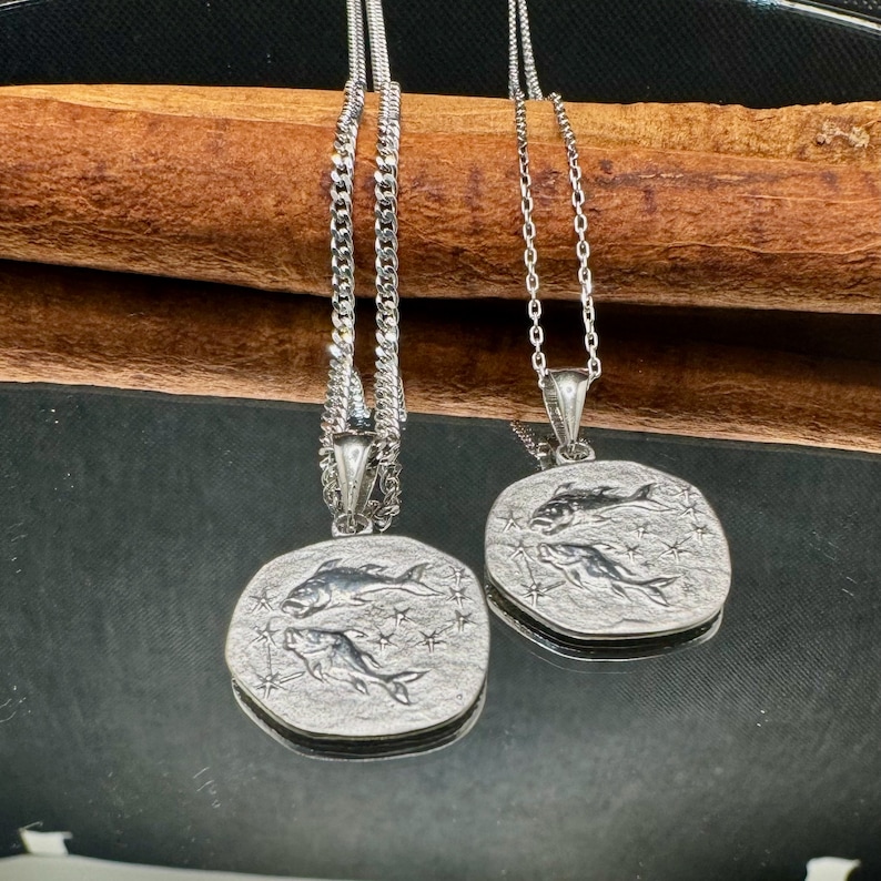 Siver Pisces pendant in the shape of an old coin on a mirror backround. The Pisces symbol is embossed and shiny compared to the background. The ground is a matte color.