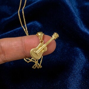 Op de afbeelding: Een goudkleurige metalen hanger in de vorm van een akoestische gitaar, met een gladde body en een textuurrijke hals met stempennen. De hanger is bevestigd aan een fijne goudkleurige ketting, waardoor een ketting ontstaat die aan een metalen oogje hangt.