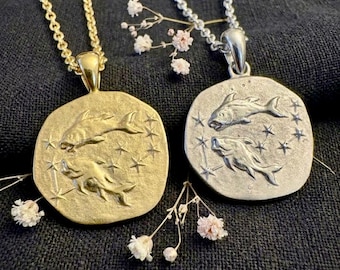 May include: Two pendant necklaces, one gold-toned and one silver-toned, each featuring a circular charm with two fish and star accents. The gold necklace has a matching chain, while the silver one has a chain with small, round links. Delicate white and pink flowers are placed around the necklaces.