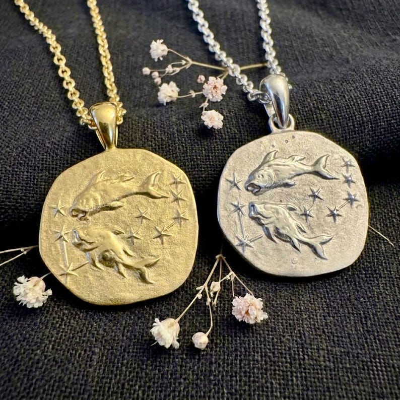 Puede incluir: Dos collares con colgantes, uno dorado y otro plateado, cada uno con un dije circular con dos peces y detalles de estrellas. El collar dorado tiene una cadena a juego, mientras que el plateado tiene una cadena con eslabones peque&ntilde;os y redondos. Delicadas flores blancas y rosas adornan los collares.