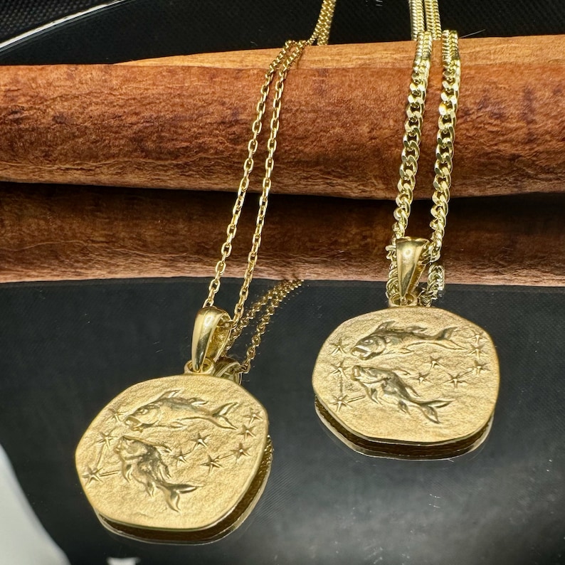 Gold Pisces pendant in the shape of an old coin on a gray plain background. The Pisces symbol is embossed and shiny compared to the background. The ground is a matte color.